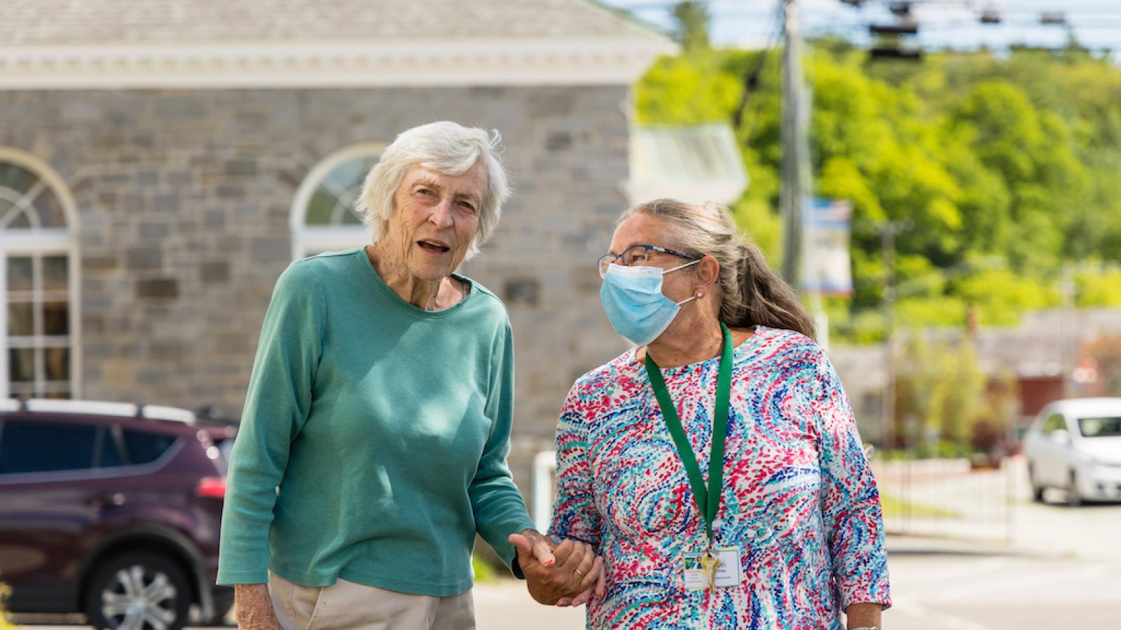 A senior member with a skilled nurse, holding hands outside.