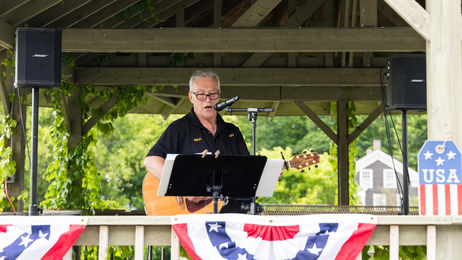A man playing an acoustic guitar outside, under a gazebo. An American flag banner is draped over the railing.
