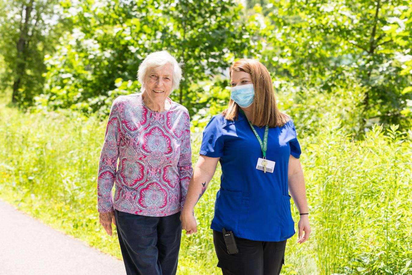 A resident and nurse walking hand in hand outside.