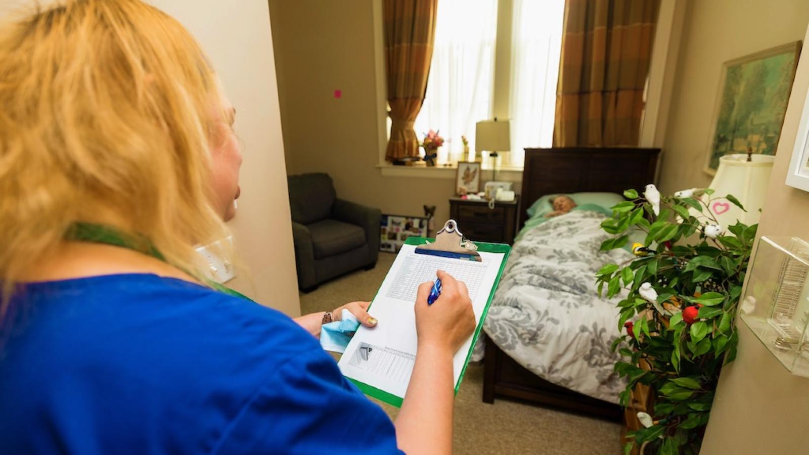 A nurse with a clipboard is checking on a resident laying in their bedroom.