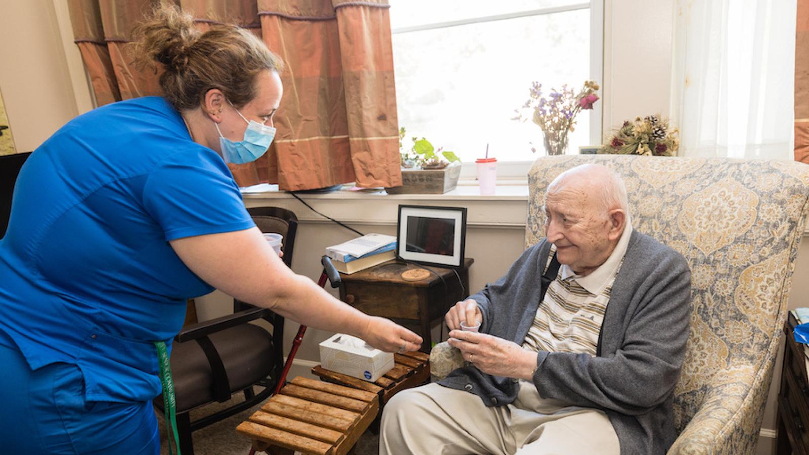 A nurse in blue scrubs giving medication to a senior resident in a recliner