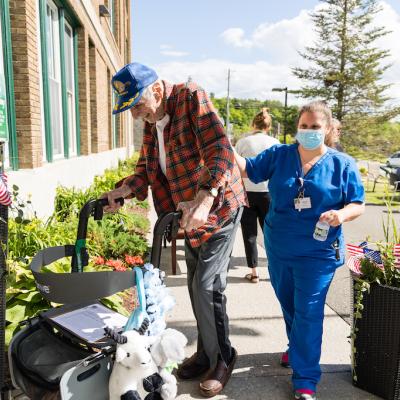 A resident using a walker to enter the facility, with help from a nurse in blue scrubs.