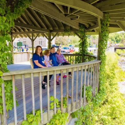 Two nurses with two residents, sitting outside under a gazebo. They are looking and the nearby river.