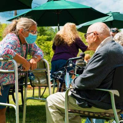 A resident sitting down as a nurse, leaning on a walker is talking to them.