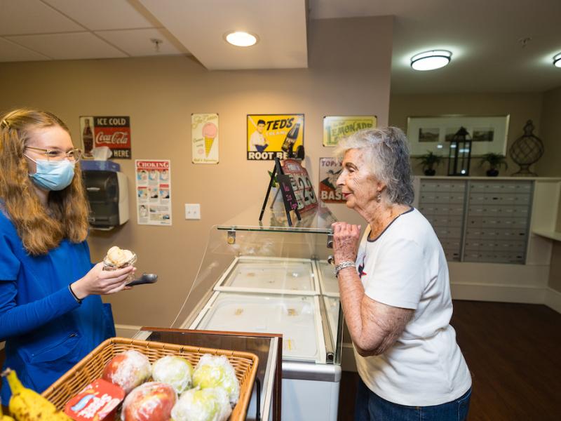 A nurse scooping ice cream into a cone, for a resident looking on.